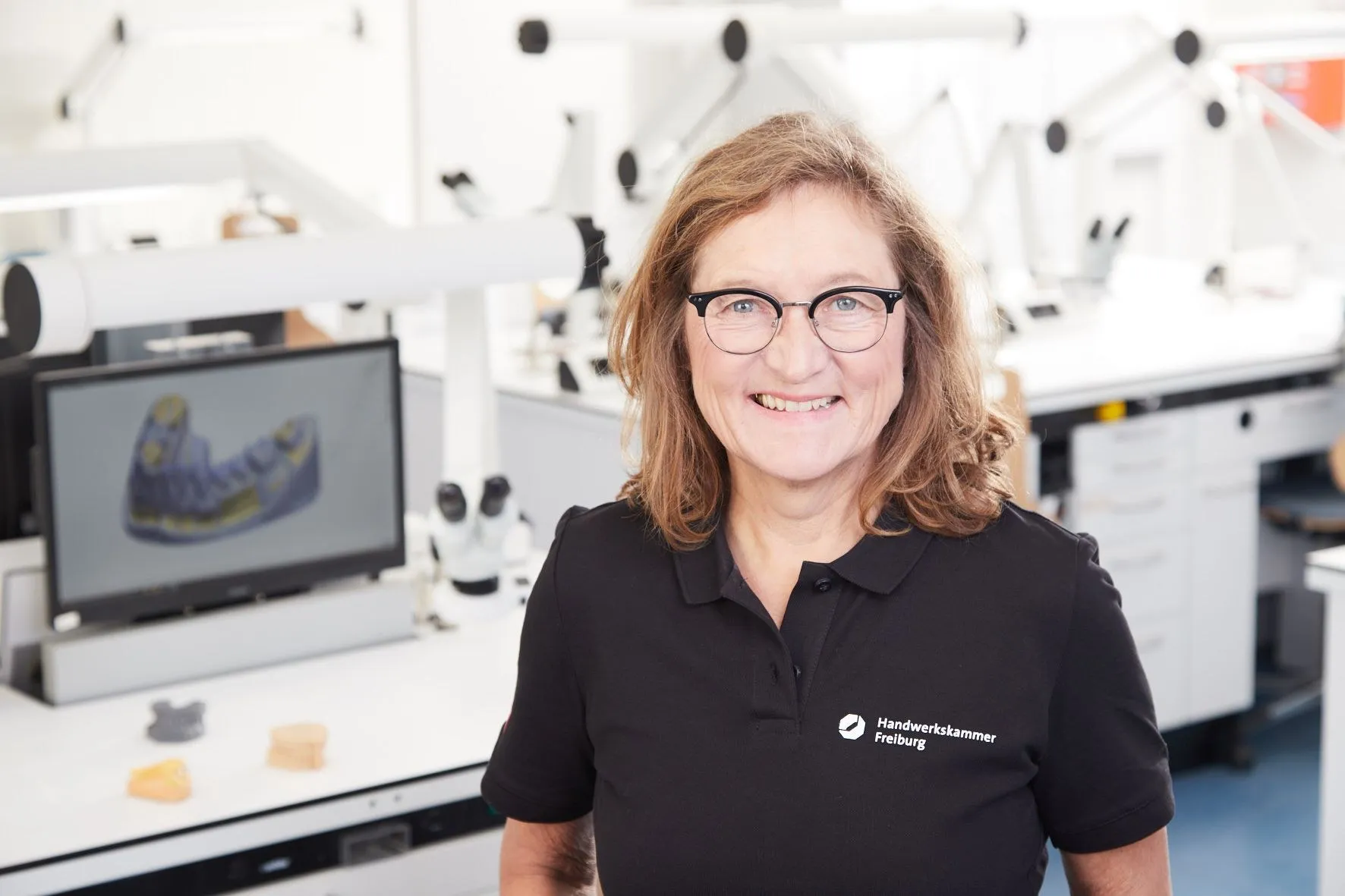 Sonja Weiss in schwarzem Poloshirt, lächelnd in einem hellen Dentallabor.